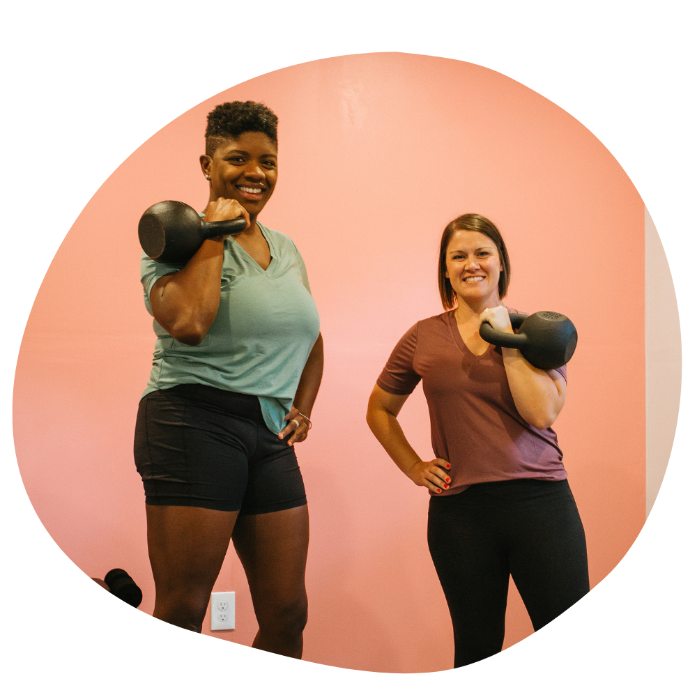 A tall black woman with short hair and a short white woman with chin-length hair hold kettle bells at their shoulders while smiling for the camera