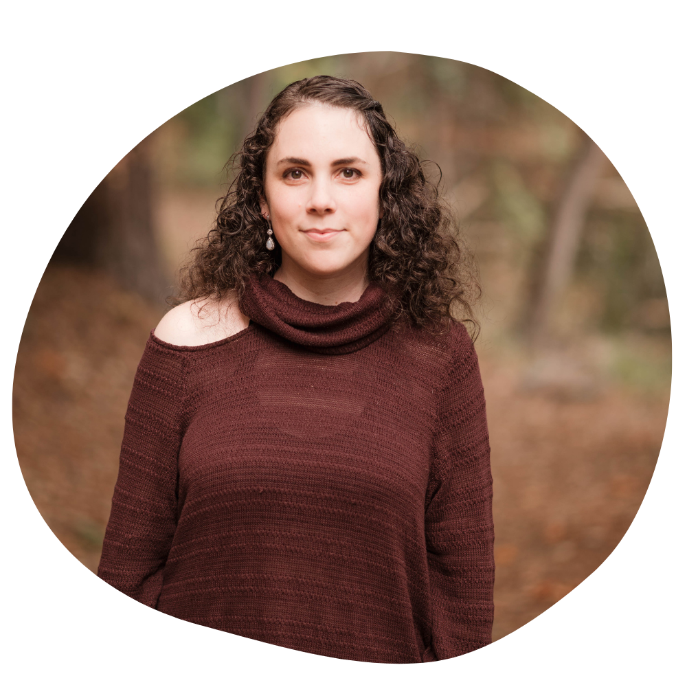 A photo of a woman with porcelain skin and curly brown hair standing in the woods and smiling softly 