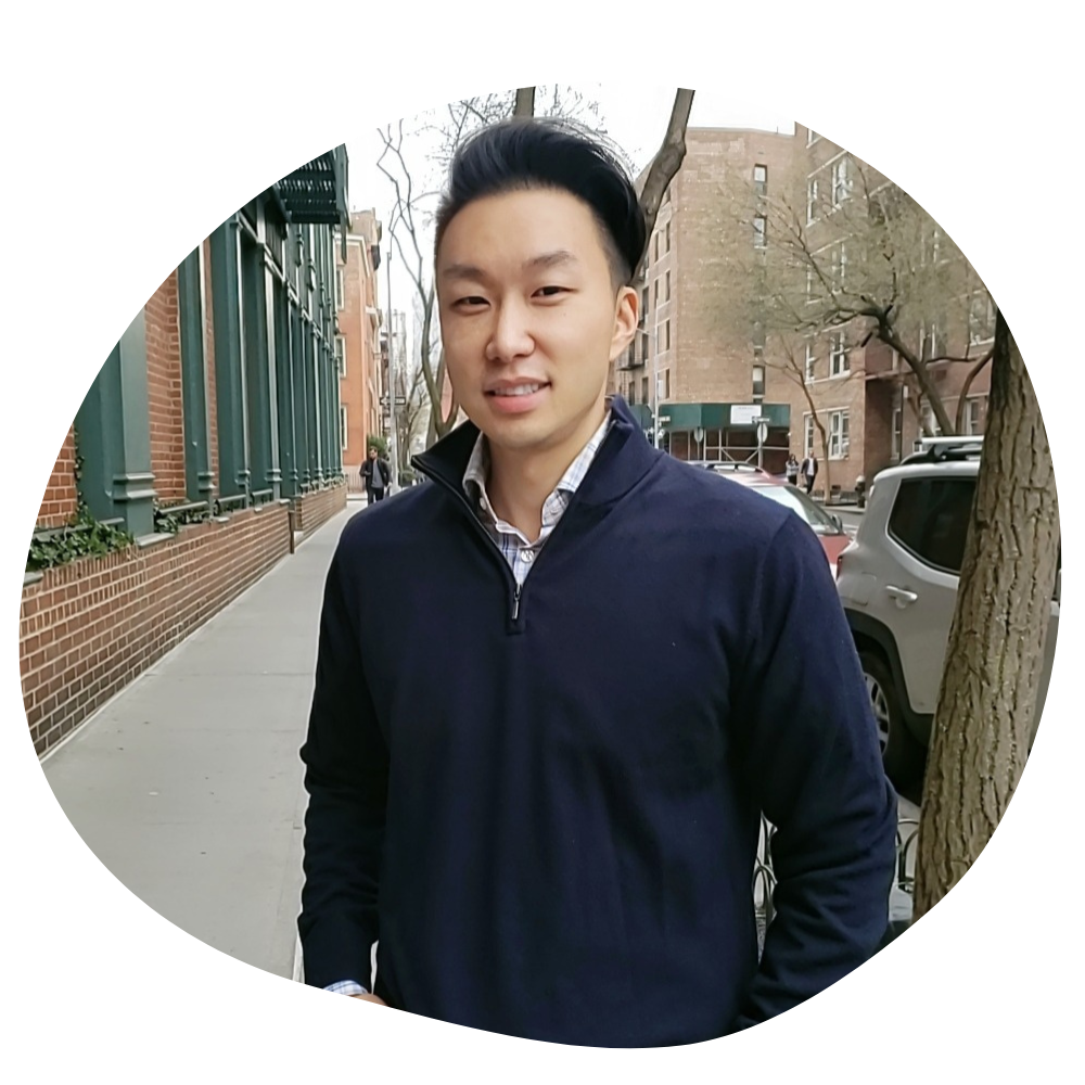 An asian man with short hair in a blue pullover stands on a city street in front of a small tree