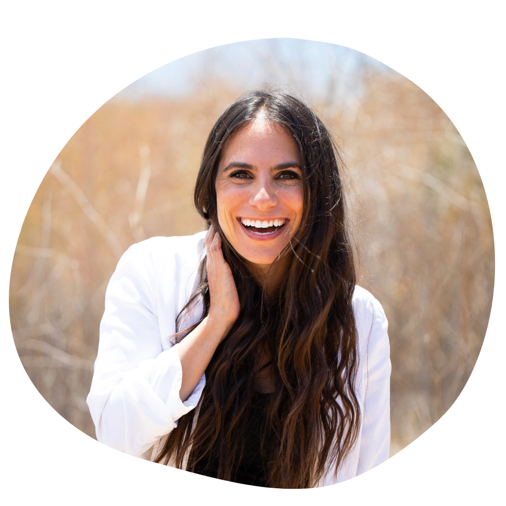 Beautiful woman with olive skin and long brown hair smiles vibrantly at the camera, her white shirt contrasting her dark hair, with a blurred cornfield behind her