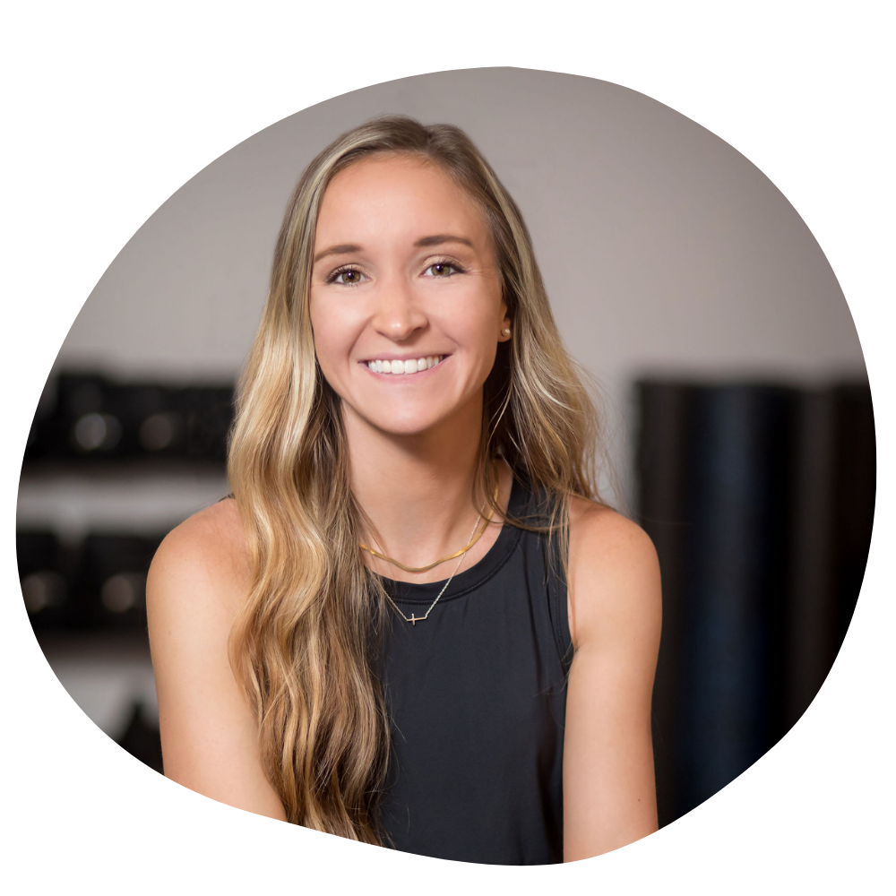 A thin woman with long wavy brown hair sits in a fitness studio and smiles for the camera