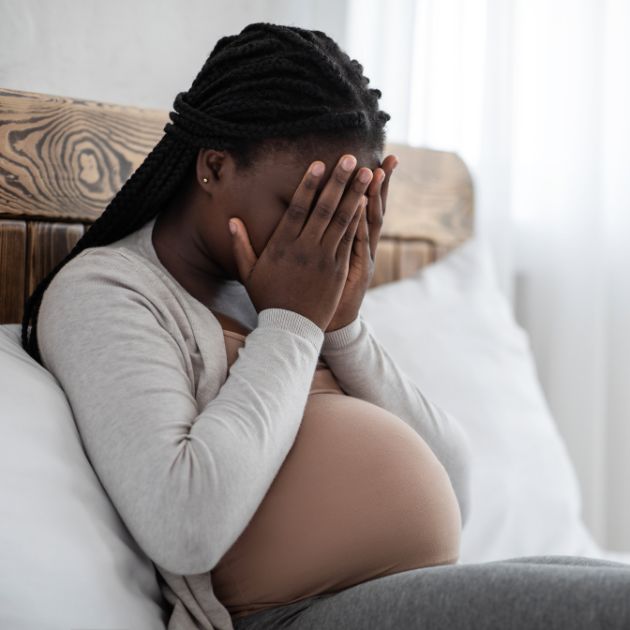 An African American woman with her hair pulled half back sits on a wooden bed with white linens, her pregnant belly visible, with her hands covering her face as if she's frustrated