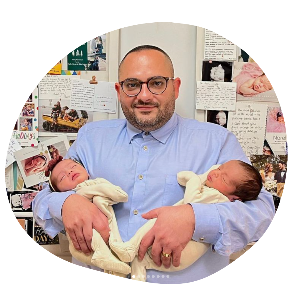 a man with buzzed black hair and a scruffy beard wears camel colored glasses while holding two babies