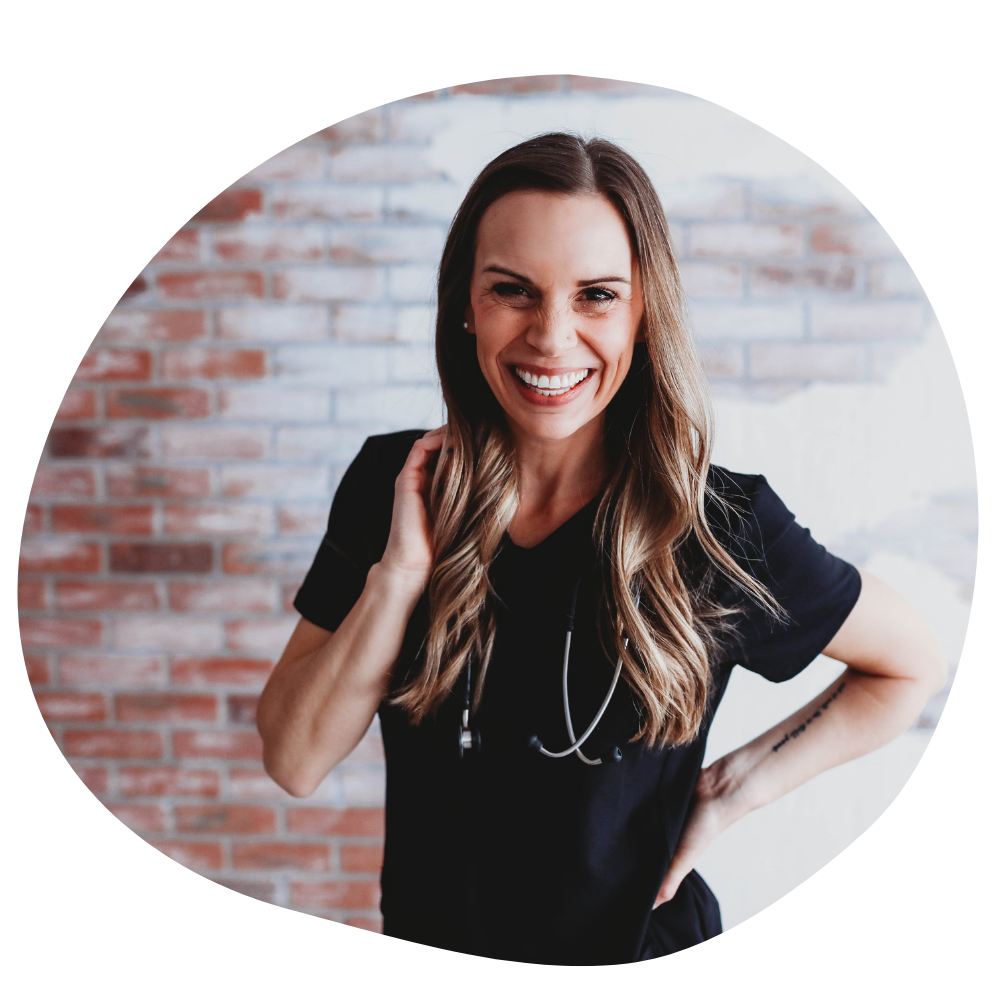 Thing nurse with long light brown hair wears black scrubs with a stethoscope around her neck while smiling for the camera in front of a whitewashed brick wall