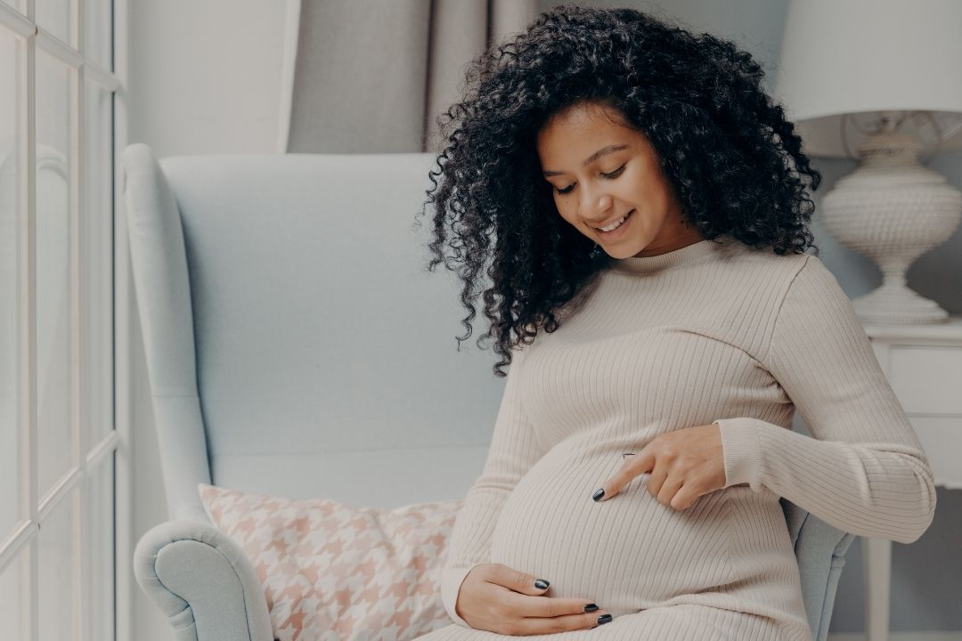 a beautiful African American woman with vibrant curly hair sits in a light blue chair wearing a tan dress, smiling at her pregnant belly, which she points to with her freshly polished finger